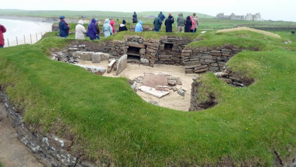 One of the dwellings at Skara Brae with Skaill House in the background and the Bay of Skaill to he left.